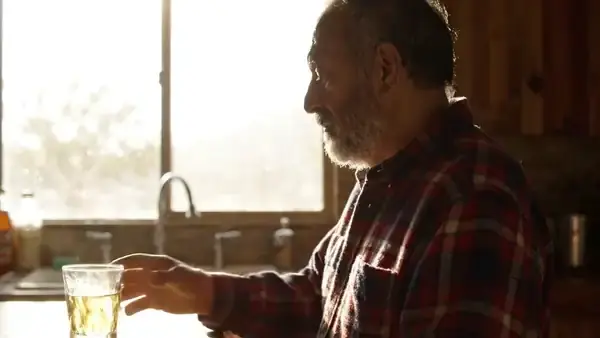 An older man in a flannel shirt taking a slow sip from a glass of warm water at a kitchen window, seen in profile